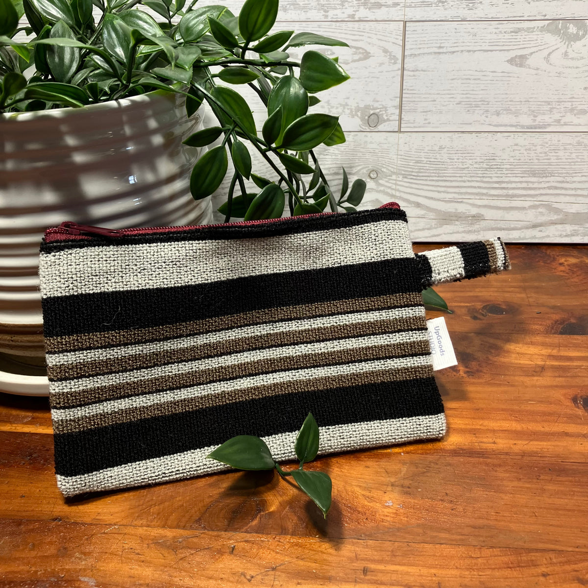 A woven zip pouch with horizontal stripes in black, beige, and brown, resting on a wooden tabletop next to a potted plant.