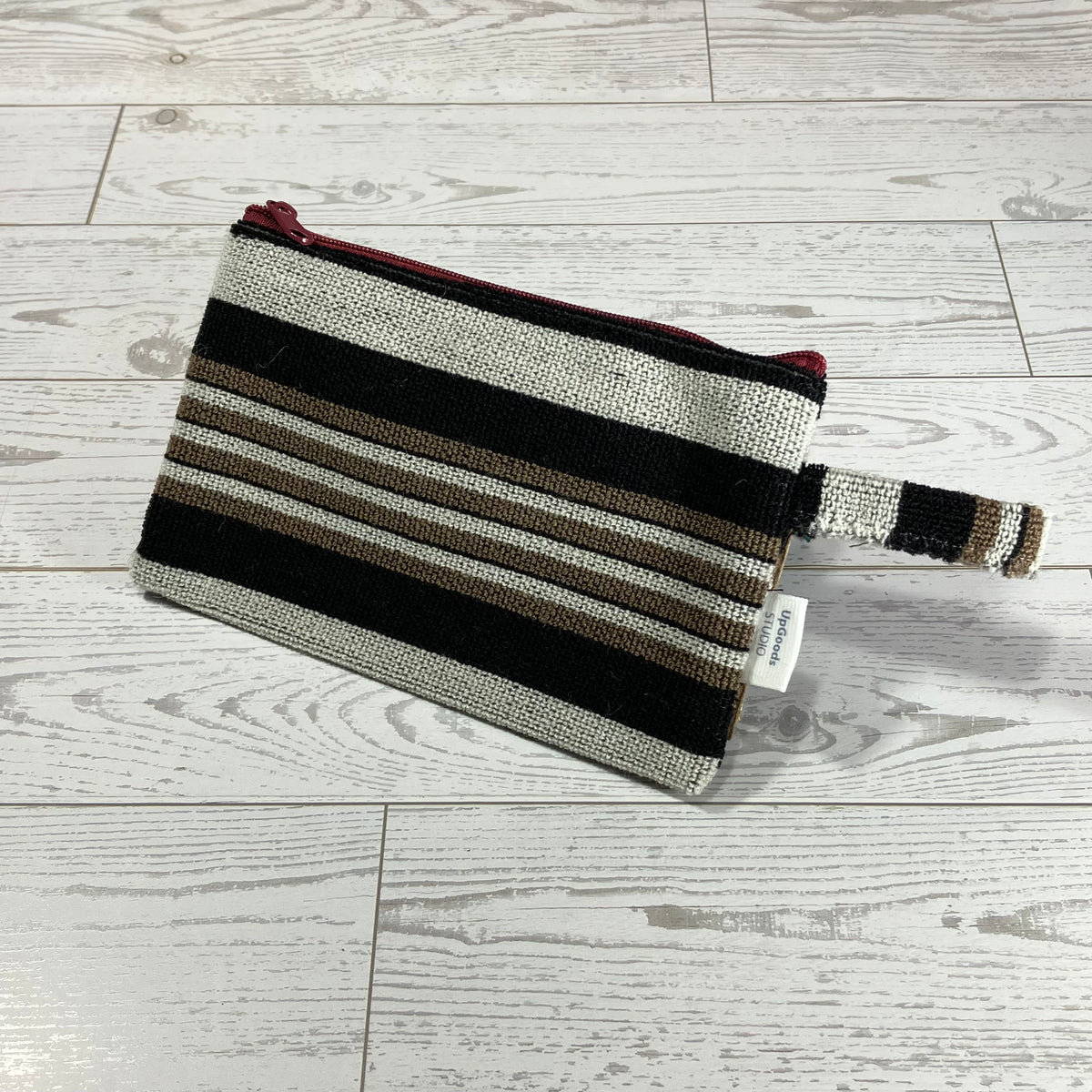 A woven zip pouch with horizontal stripes in black, beige, brown, and white, featuring a red zipper and a matching woven wrist strap, placed on a light gray wood plank floor.