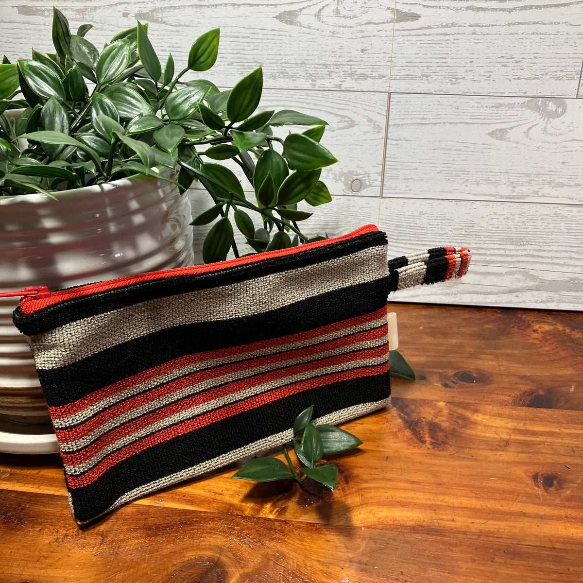 A woven zip pouch with horizontal stripes in black, beige, and red, resting on a wooden table next to a potted green plant.