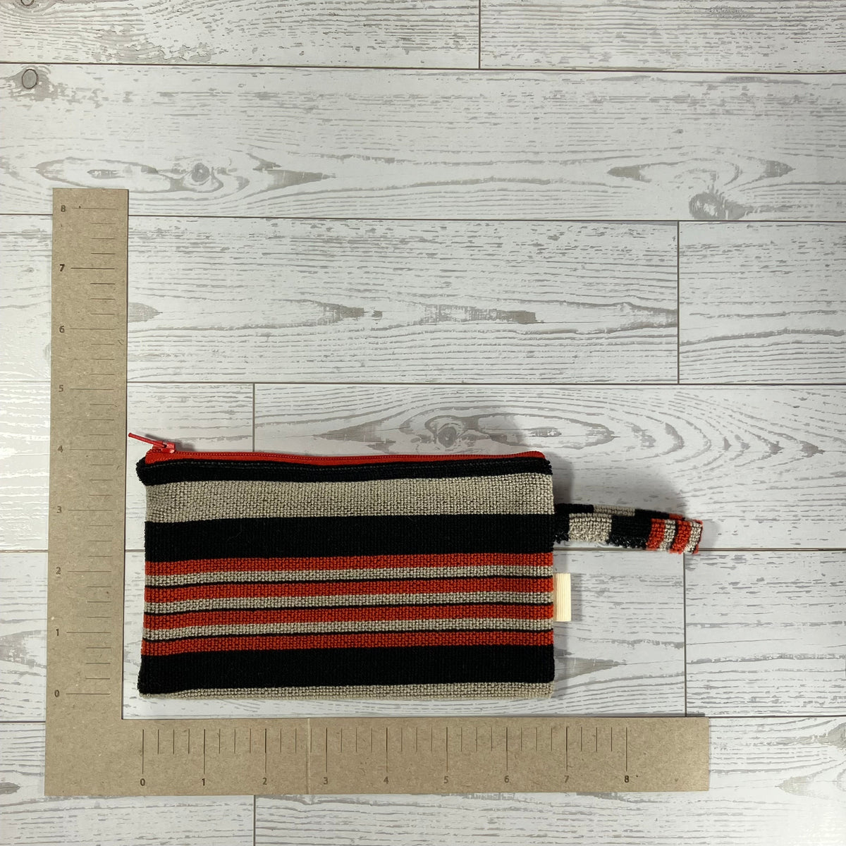A striped woven zippered pouch with a top handle, shown on a light gray wood-plank floor with cardboard rulers for scale.