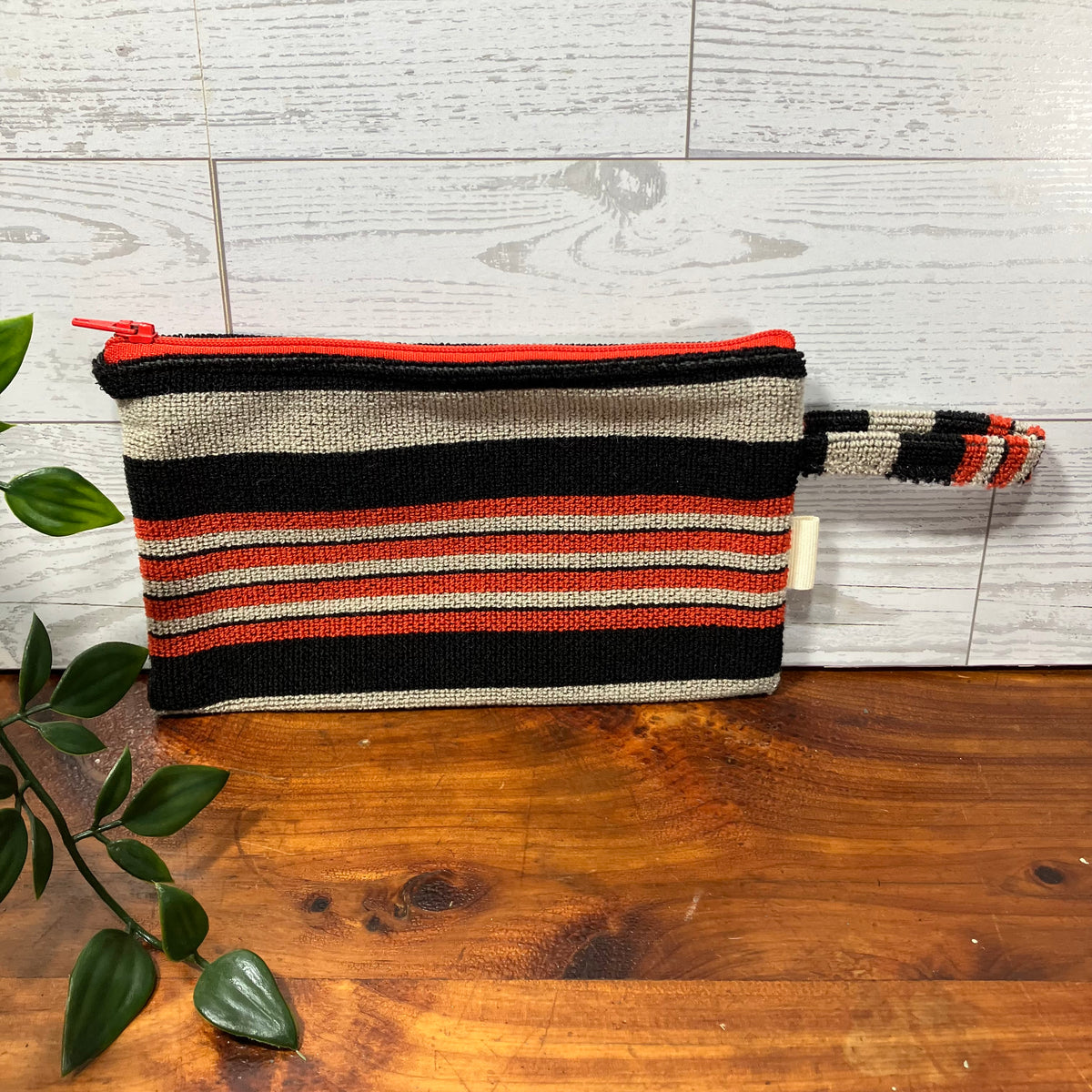 A woven zip pouch with horizontal stripes in black, beige, and rust red, resting on a wooden surface against a white wood paneled wall.