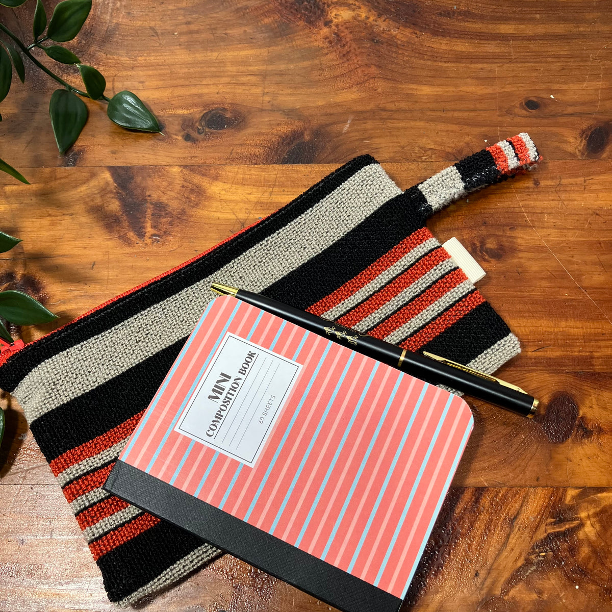 A striped woven pouch with a matching zipper pull, styled on a wooden tabletop alongside a pink striped notebook and a black pen.