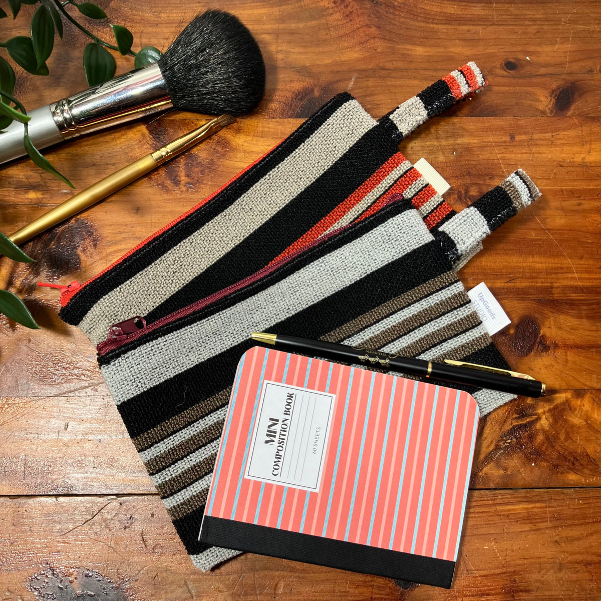 A striped woven zip pouch laid on a wooden tabletop, styled with a coral-and-white striped notebook, a black pen, a makeup brush, and a gold pencil. No person or animal is present, so this is Product/Packaging rather than Lifestyle, even though it uses a styled flat-lay arrangement.