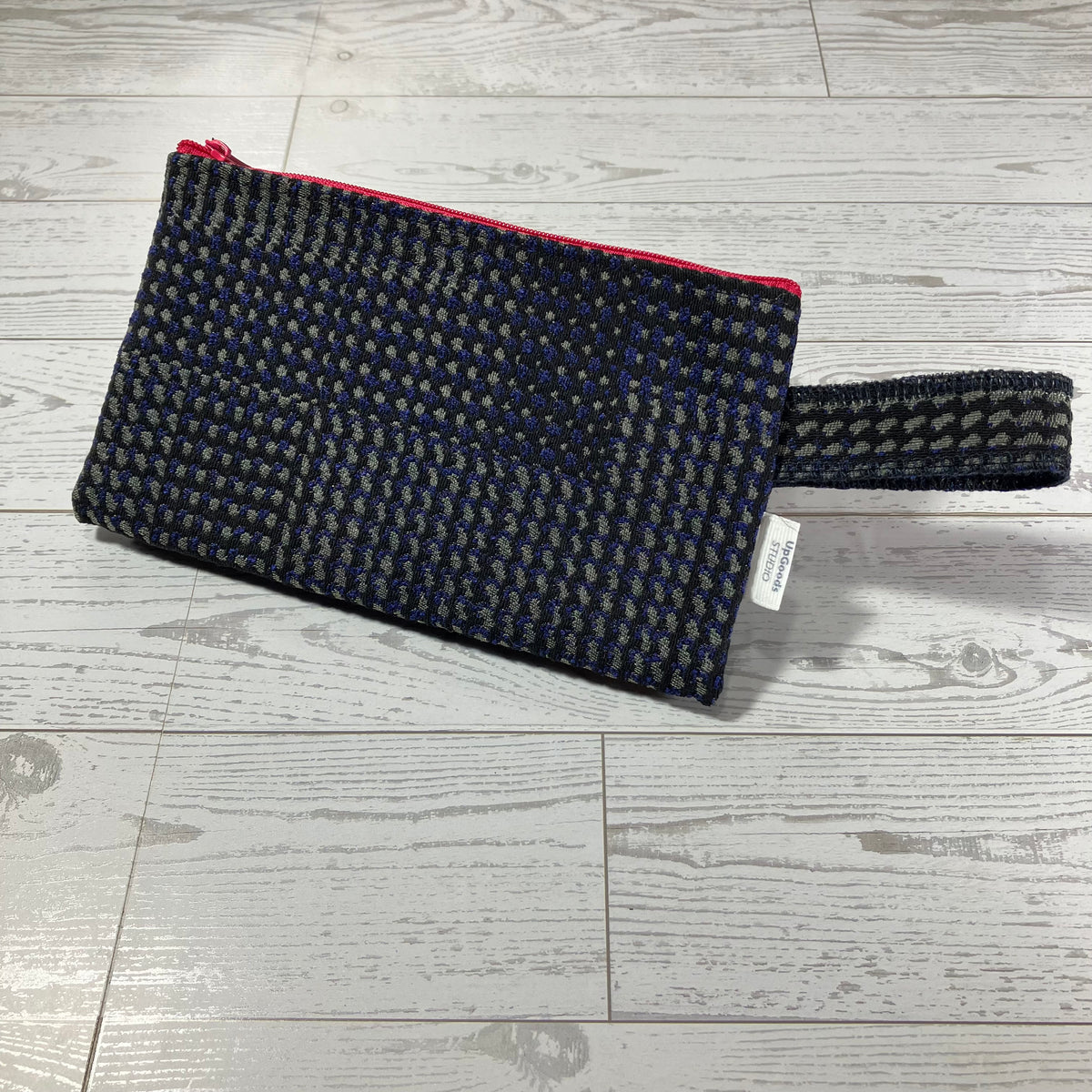 A woven clutch pouch with a black and gray patterned body, a bright pink zipper trim, and a matching wrist strap, shown on a light gray wood floor.