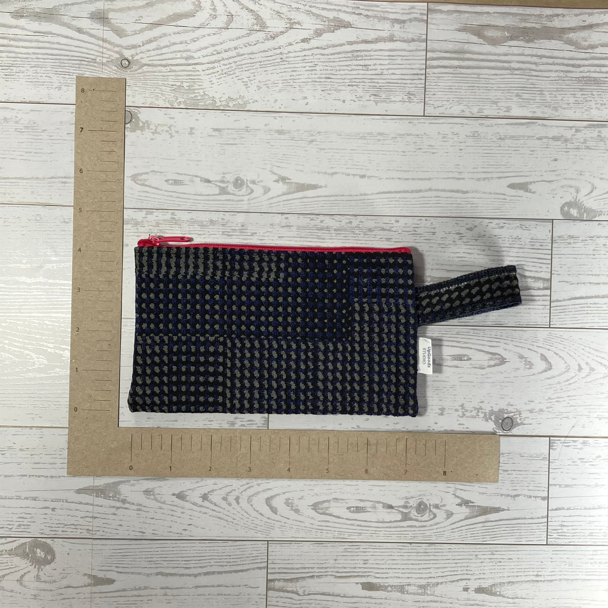A black and gray houndstooth patterned fabric pouch or bag with a red inner lining, shown on a light gray wood plank background with a cardboard L-square ruler for scale.