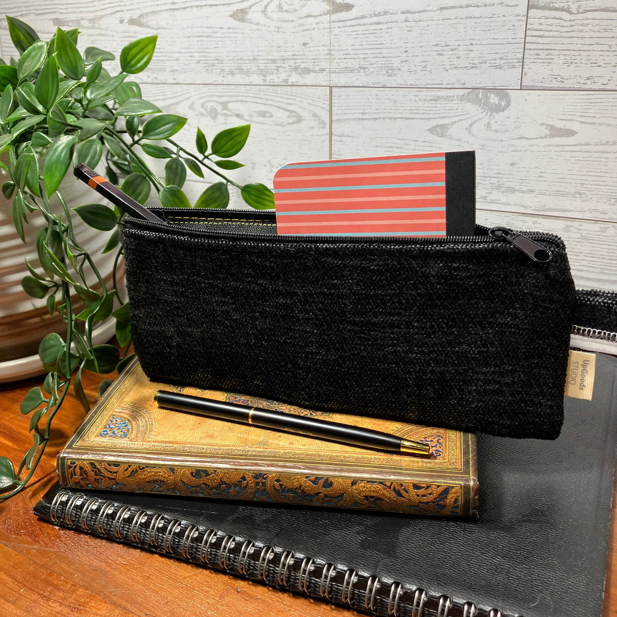 A black woven pencil pouch with a zipper closure, styled on top of stacked books with a pen resting across the front. A small striped card is partially visible inside the pouch, with a green plant and a light wood-paneled wall in the background.