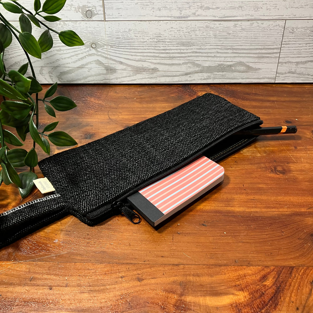 A black woven zippered pouch or belt bag lies open on a wooden tabletop, with a pink striped notebook partially tucked inside and a black pen with an orange tip resting at the zipper. A leafy plant and a light wood paneled wall are visible in the background.