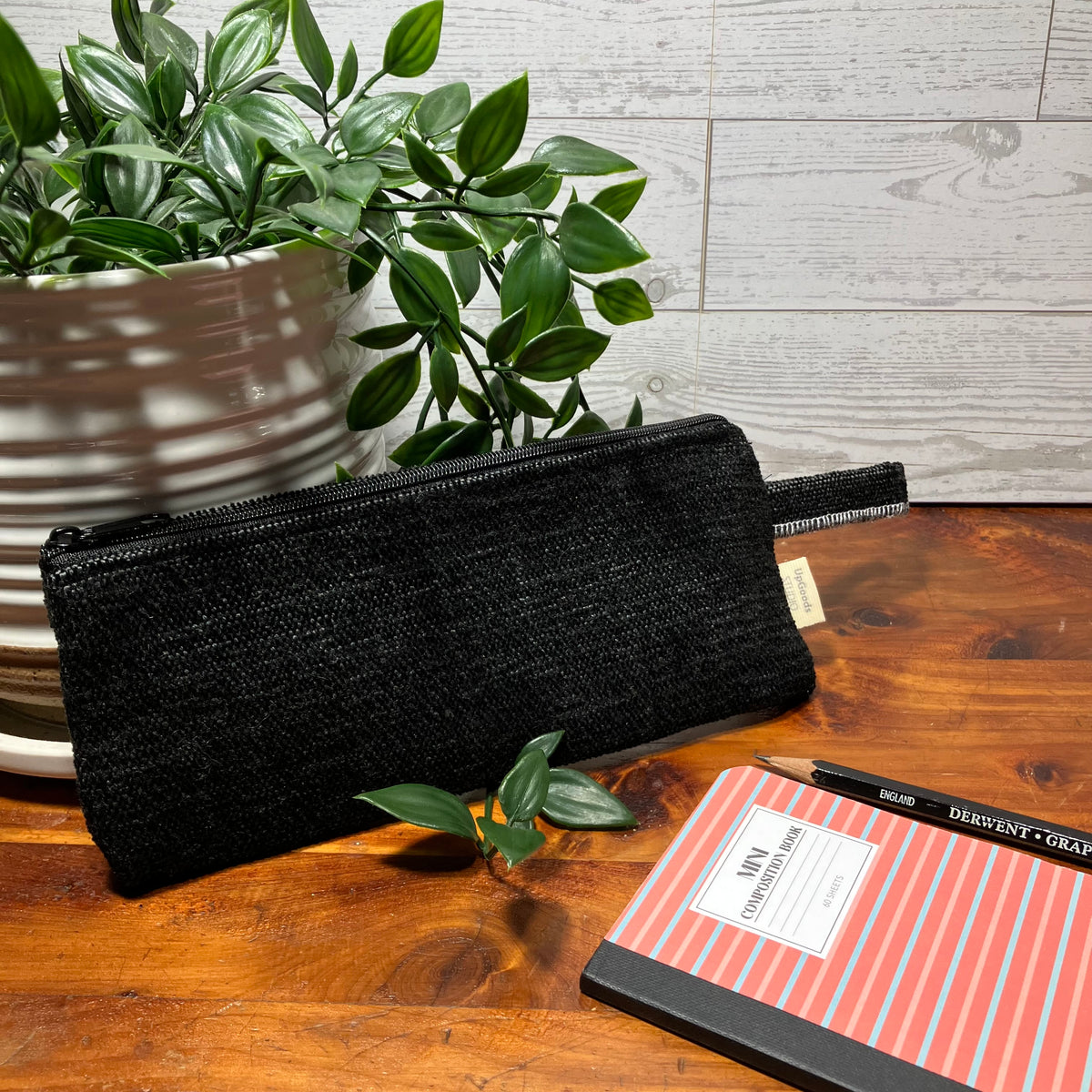 A black woven zippered pencil case with a small beige fabric tag, displayed on a wooden table next to a notebook and a potted plant.