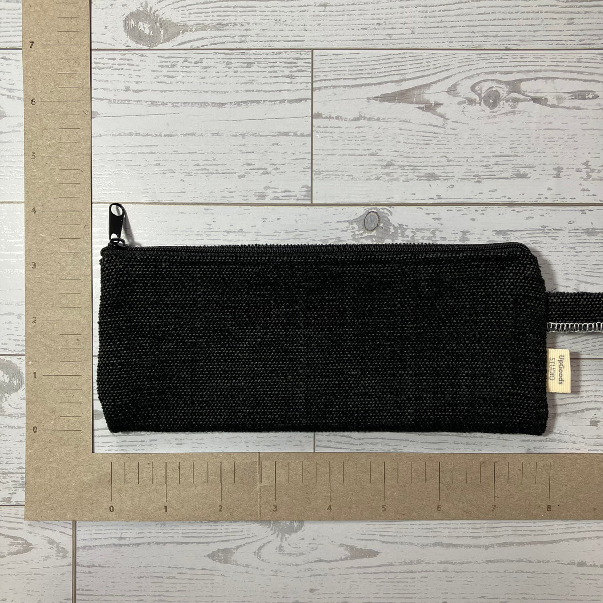 A black woven zip pouch with a small gold brand tag, laid flat on a light wood background with a cardboard ruler for scale.