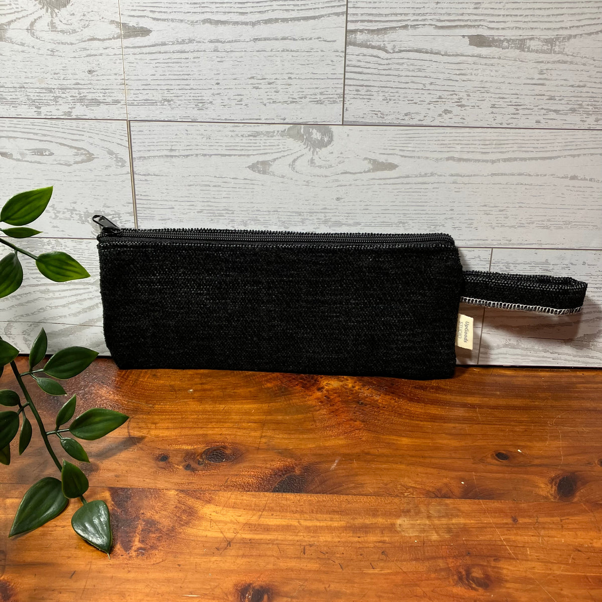 A black woven zippered pouch with a matching wrist strap, resting on a wooden surface against a light gray wood-paneled background.
