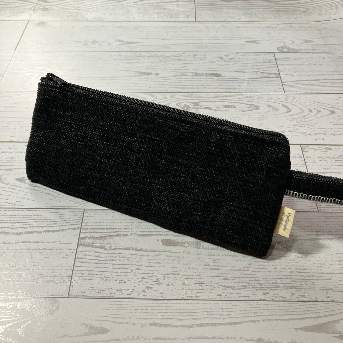 A black textured zippered pouch with a small beige fabric tag, placed diagonally on a light gray wood-plank floor.