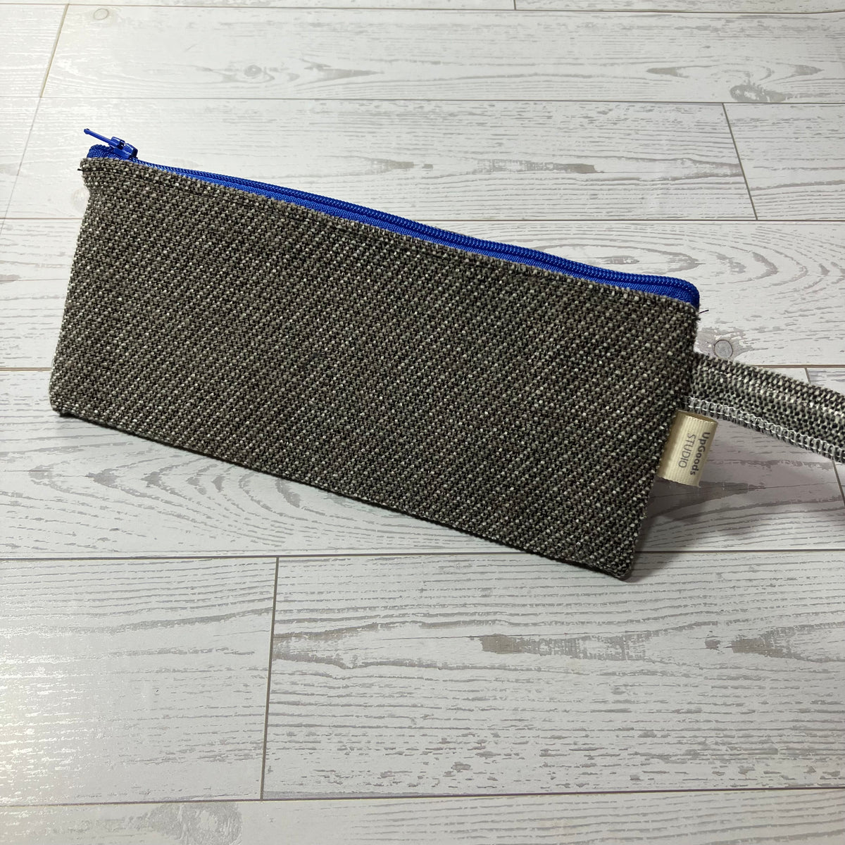 A woven rectangular pouch with a blue zipper and a matching fabric wrist strap, placed on a light gray wood plank surface.