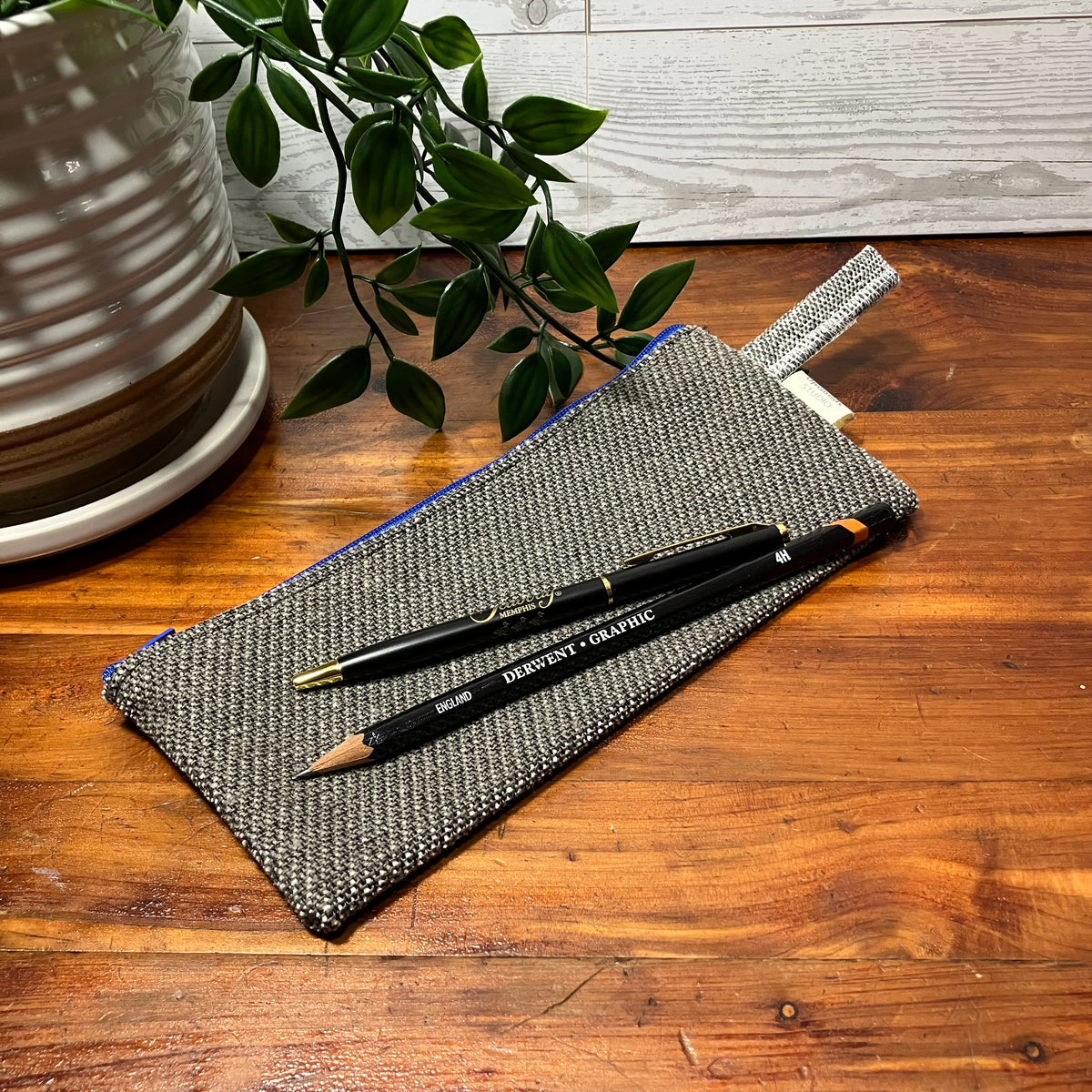 A woven gray pencil pouch with a blue zipper edge, placed on a wooden table with two black pencils resting on top. A ceramic planter and green leaves are visible in the background.