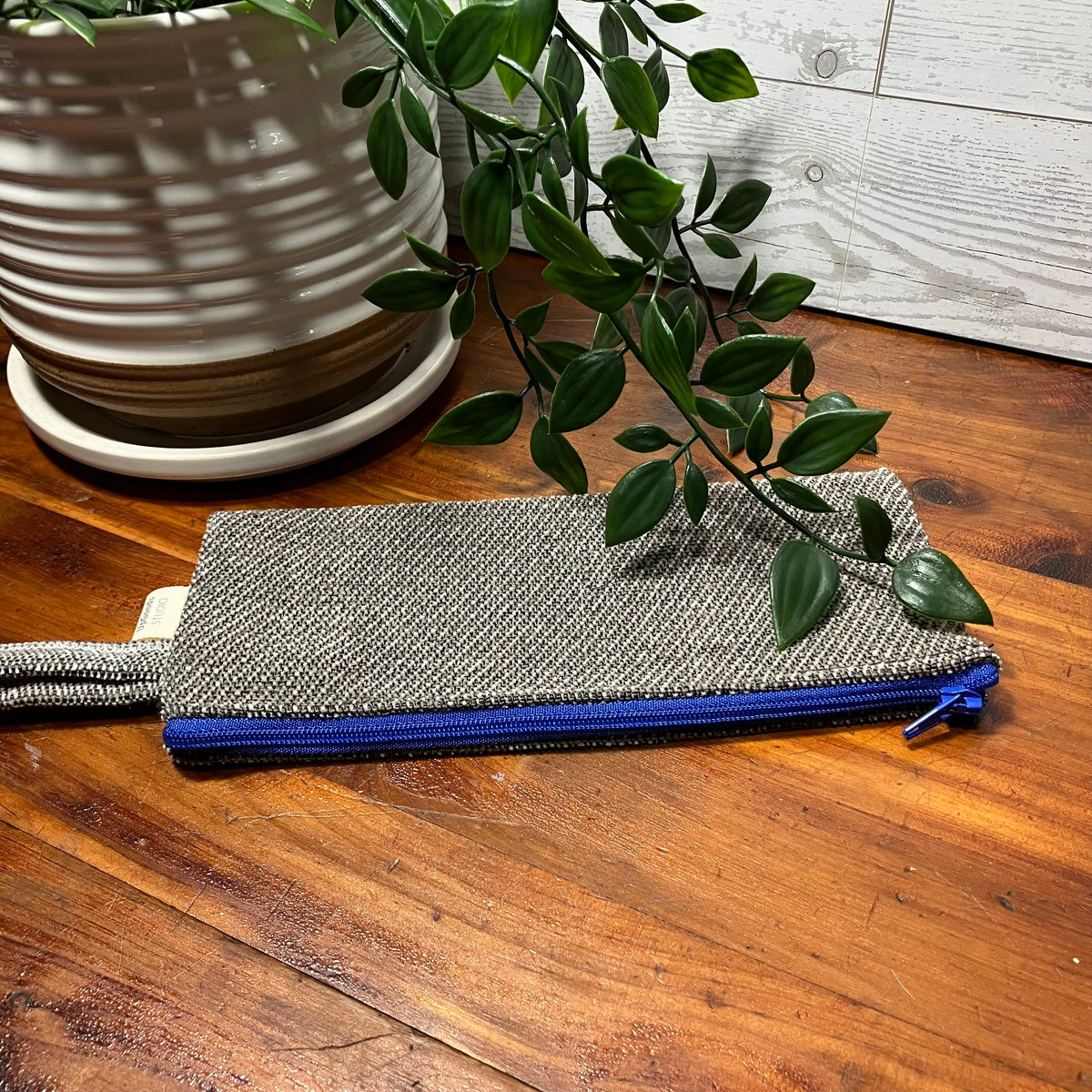 A woven fabric zip pouch with a blue zipper, placed on a wooden tabletop next to a potted plant.