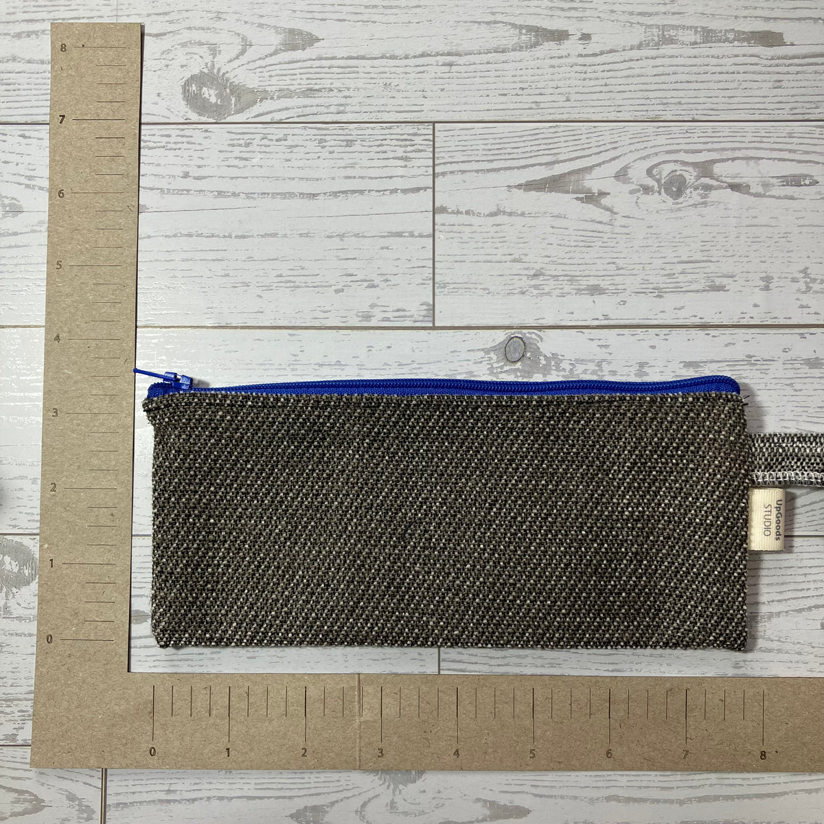 A small zippered pouch with a textured gray and black woven pattern and a bright blue zipper, shown on a white wood plank background with a cardboard ruler for scale.
