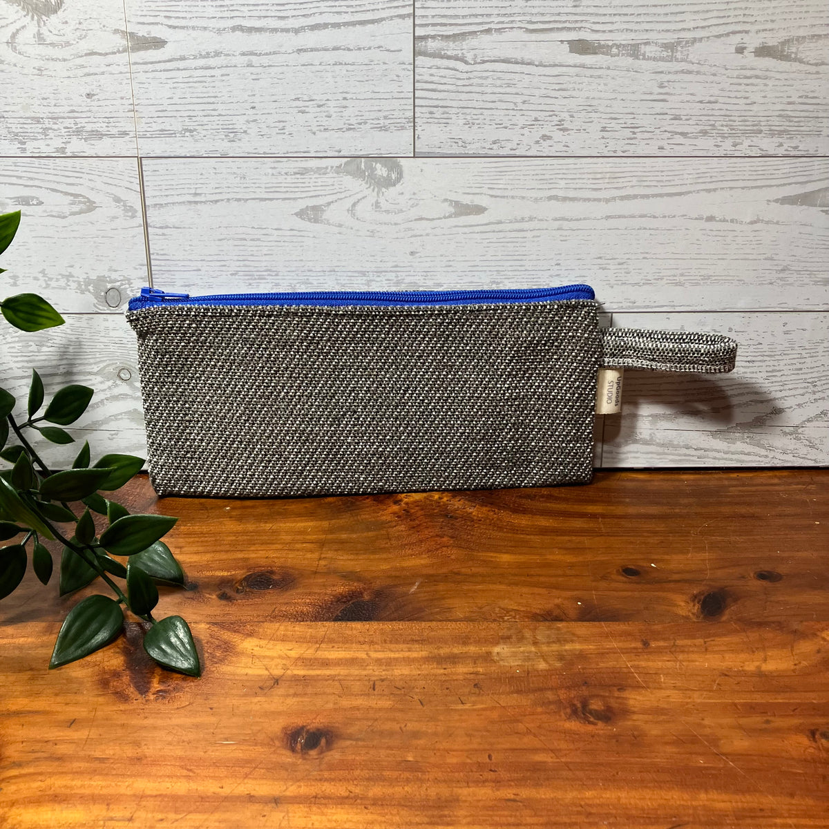 A rectangular zippered pouch in a gray and black herringbone patterned fabric, featuring a bright blue zipper and a matching fabric wristlet loop, resting on a wooden table against a light wood paneled wall.