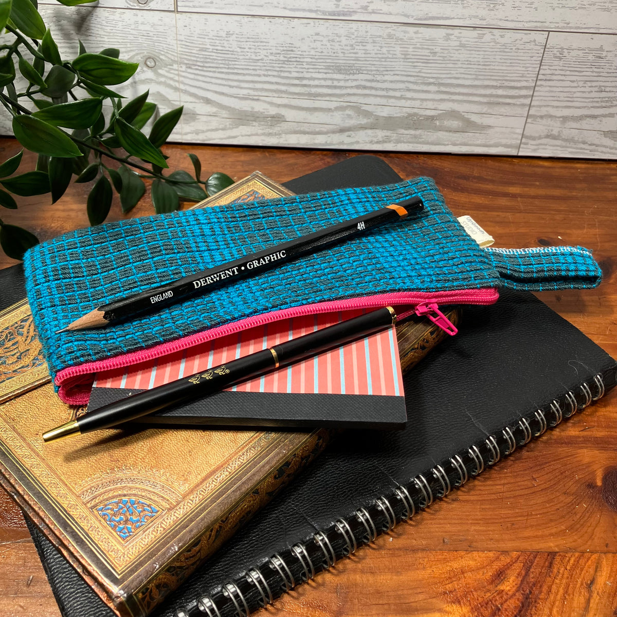 A turquoise and black woven pencil pouch with a pink zipper, styled on top of stacked books and a spiral notebook on a wooden table. A black pencil labeled 'DREISANT • GRAPHIC' rests inside the pouch.