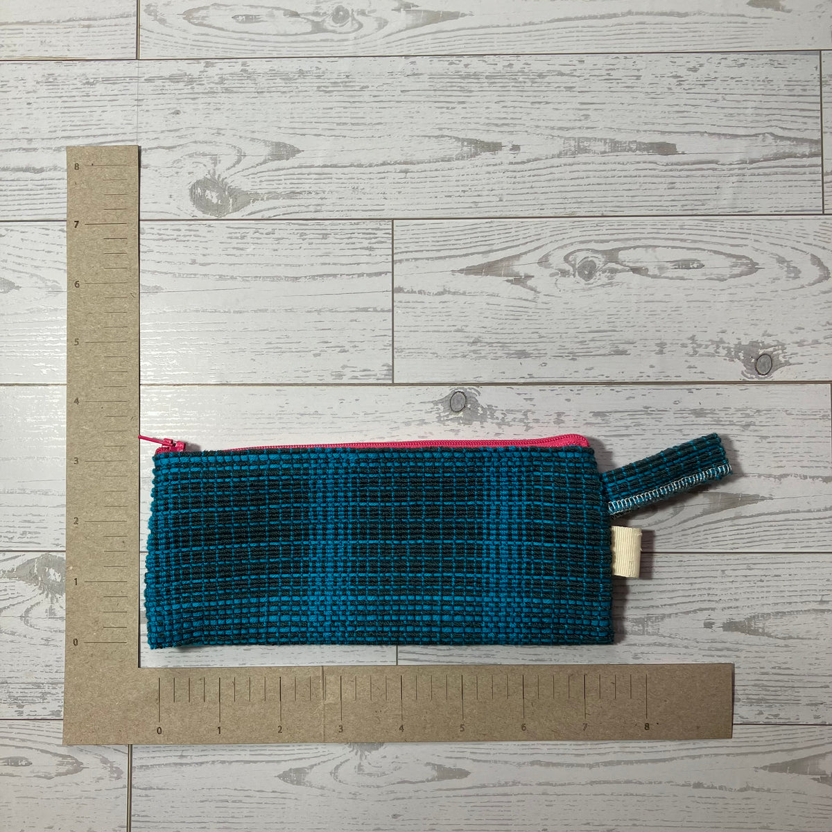 A teal and black woven zippered pouch with a pink zipper trim and a small cream tag, shown on a white wood plank background with cardboard rulers for scale.