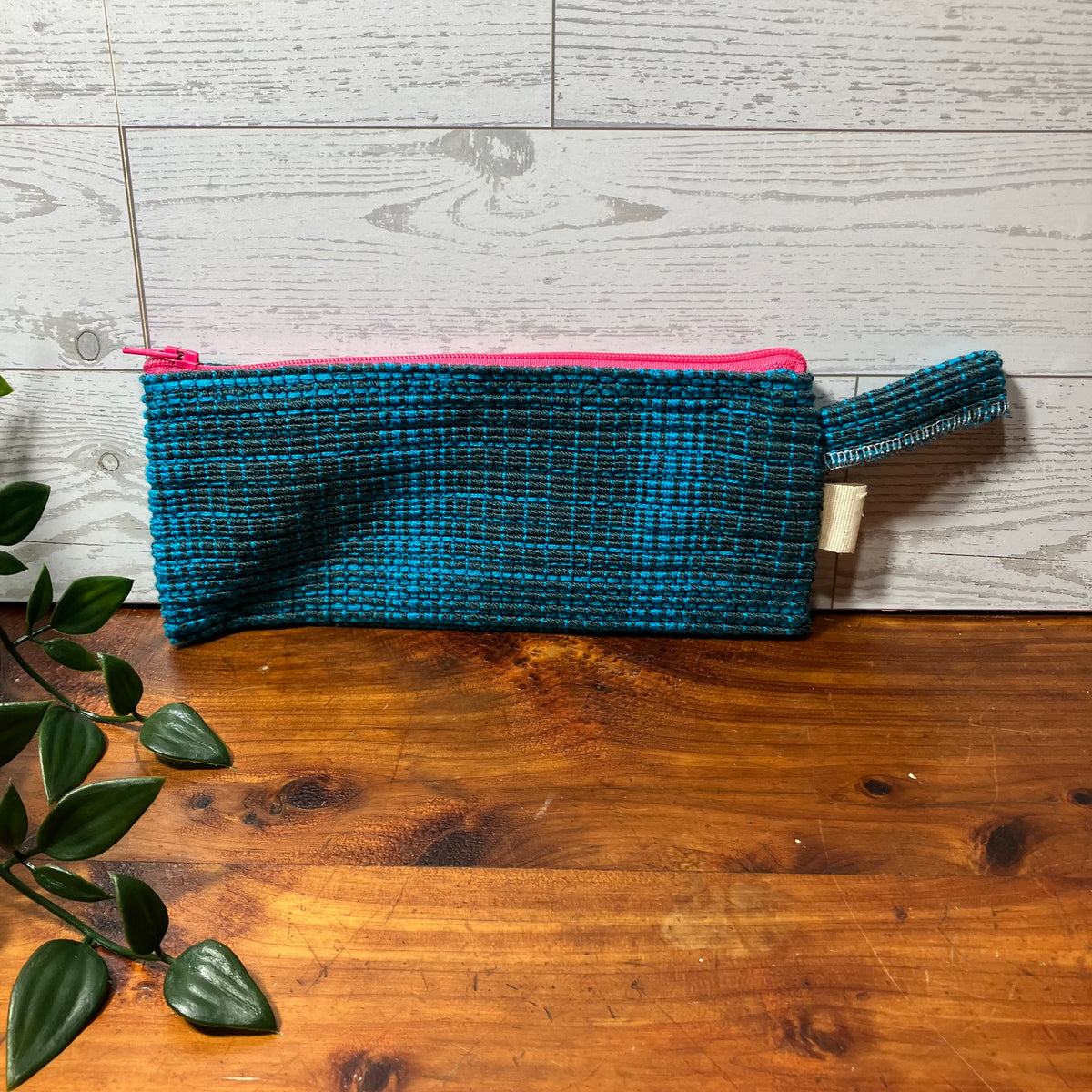 A woven teal and black cosmetic pouch with a bright pink zipper and matching pink zipper pull, resting on a wooden surface against a light gray wood plank background with a green plant leaf in the foreground.