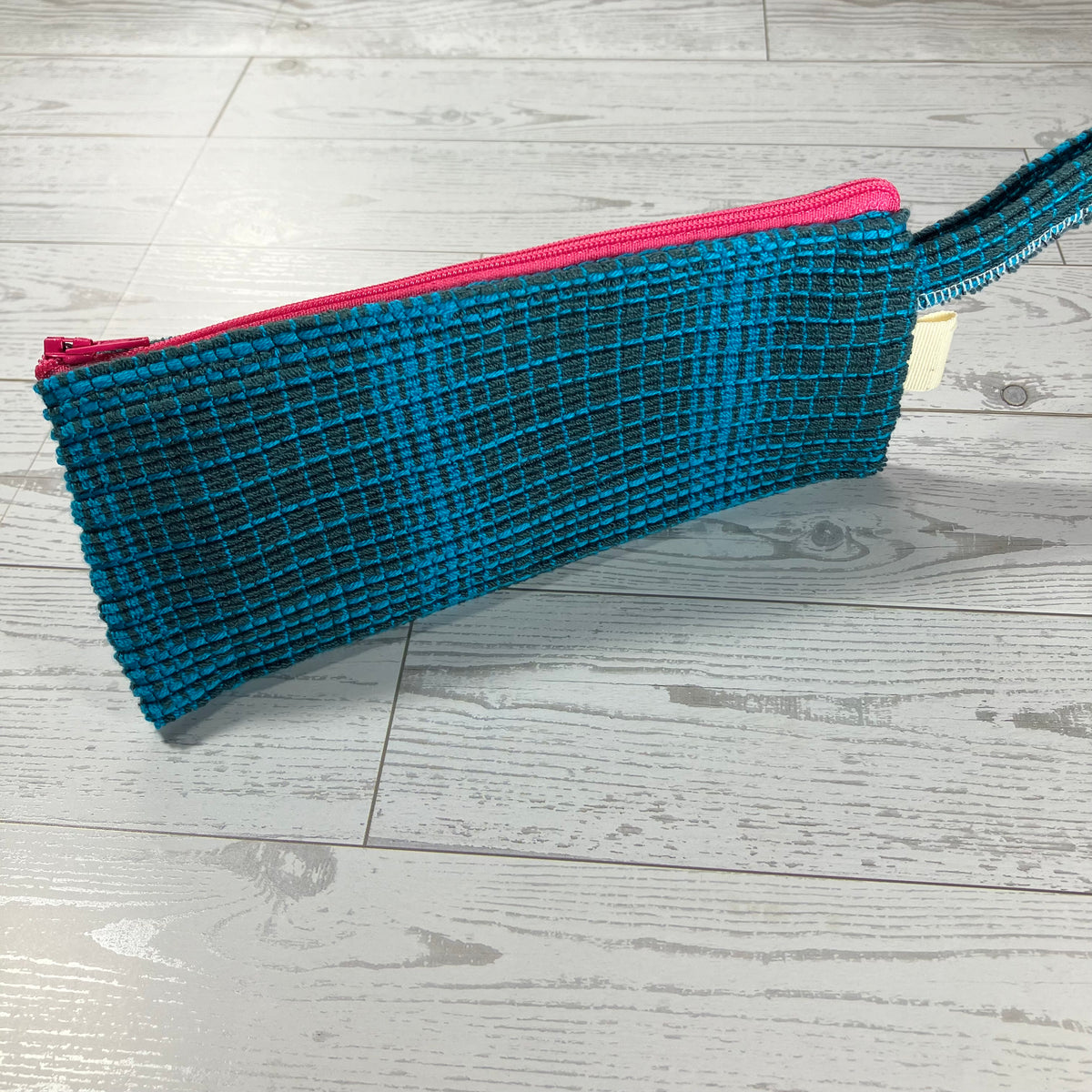 A woven clutch sized pouch bag with a textured pattern in teal and black, featuring a bright pink zipper and a matching pink wrist strap, resting on a light gray wood floor.
