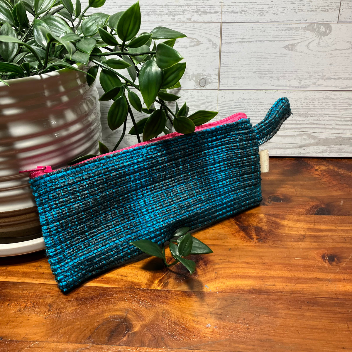 A woven teal and black zippered pouch with a pink zipper lining, styled on a wooden tabletop next to a potted green plant.