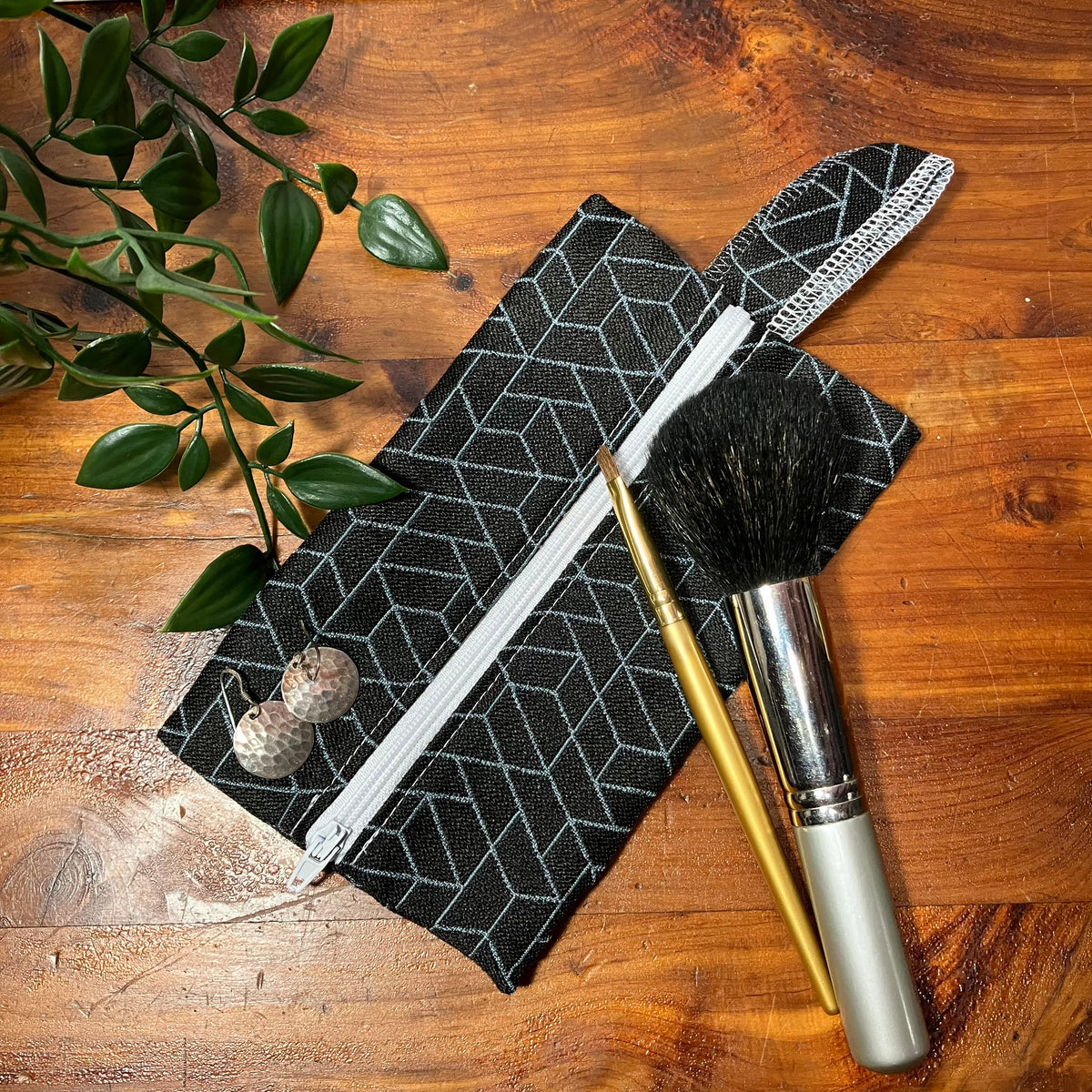 A black fabric makeup pouch with a white geometric pattern and white zipper, styled on a wooden tabletop with green leaves and two makeup brushes.
