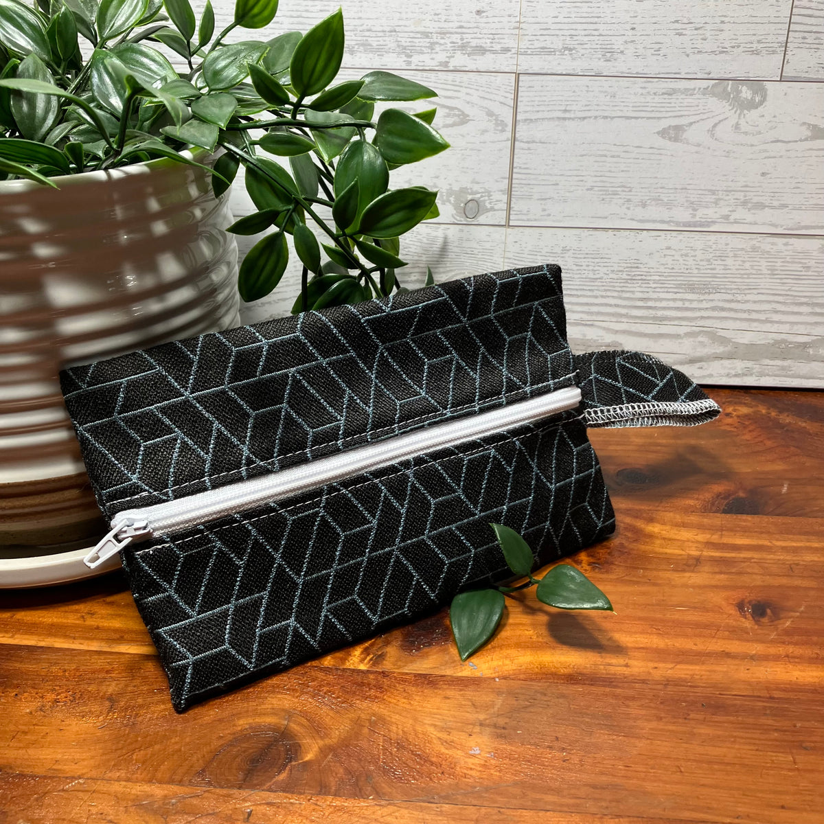 A black fabric zippered clutch or pouch with a light gray geometric line pattern, resting on a wooden tabletop next to a potted green plant.