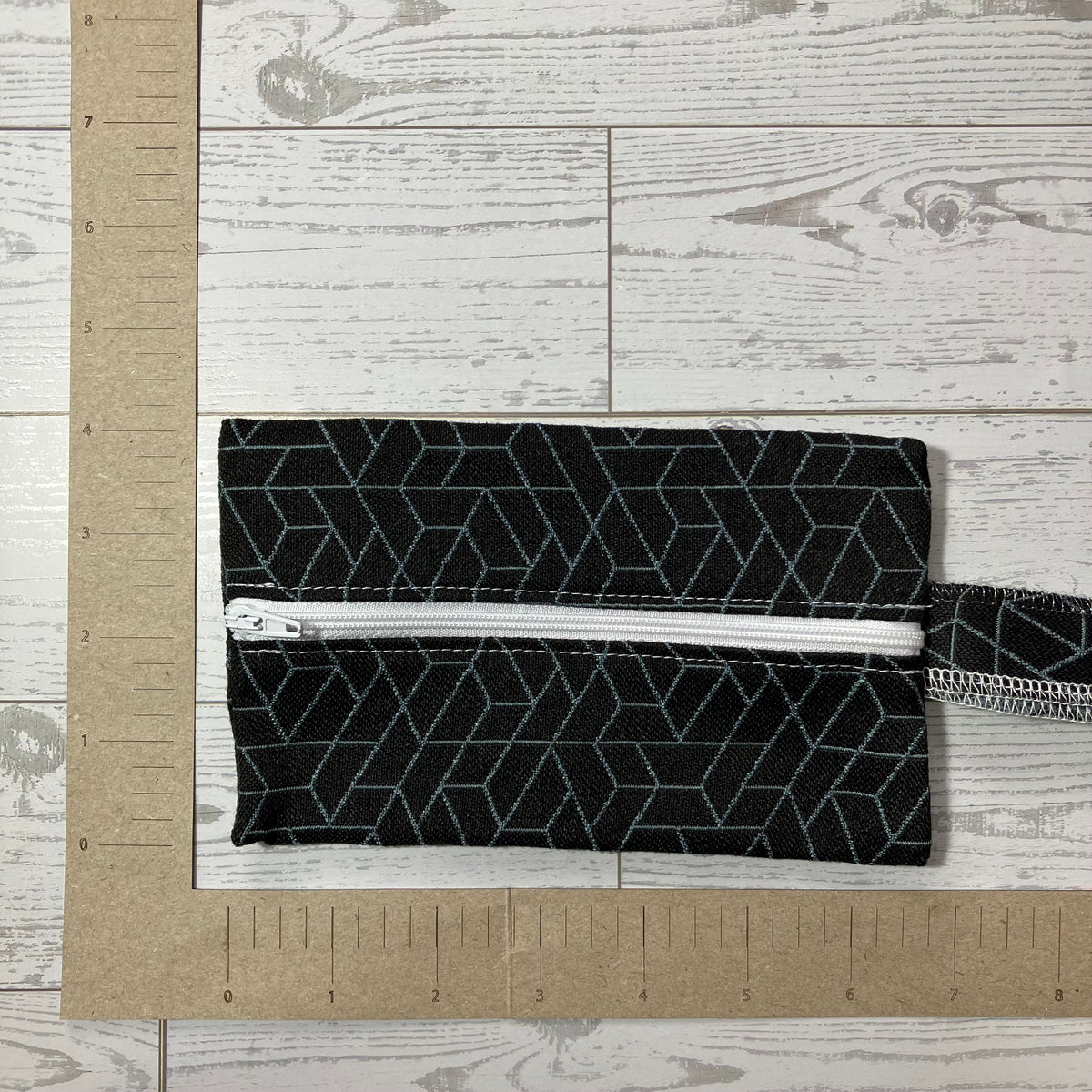 A black fabric zippered pouch with a light blue geometric pattern, shown on a white wood background with a cardboard ruler for scale.