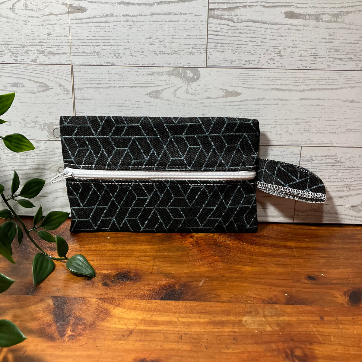 A black clutch bag with a white geometric pattern and a white zipper, resting on a wooden table against a light wood paneled wall with a green plant in the foreground.