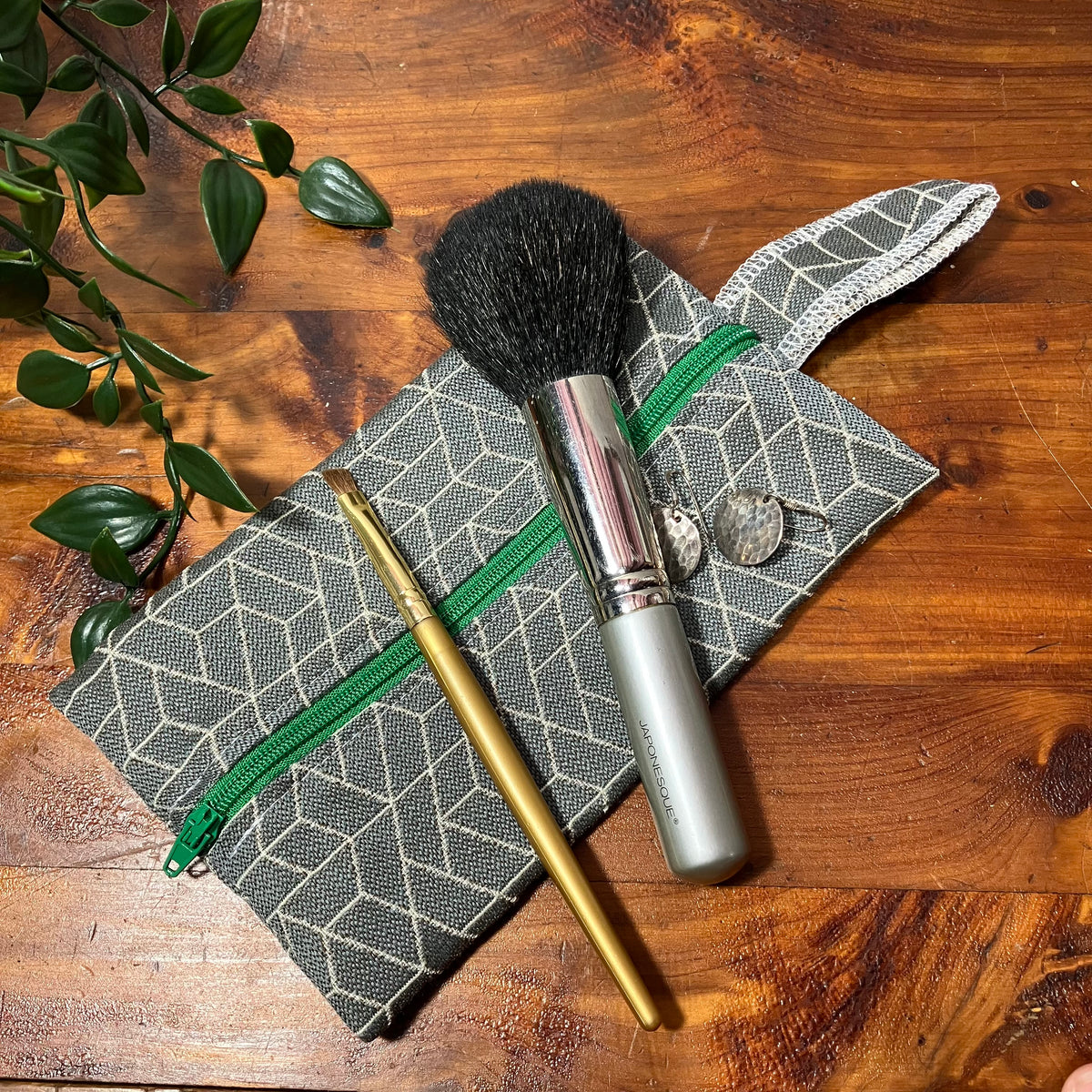 A gray and white geometric patterned makeup pouch with a green zipper, styled on a wooden table with a makeup brush, a gold crochet hook, and a small plant sprig.