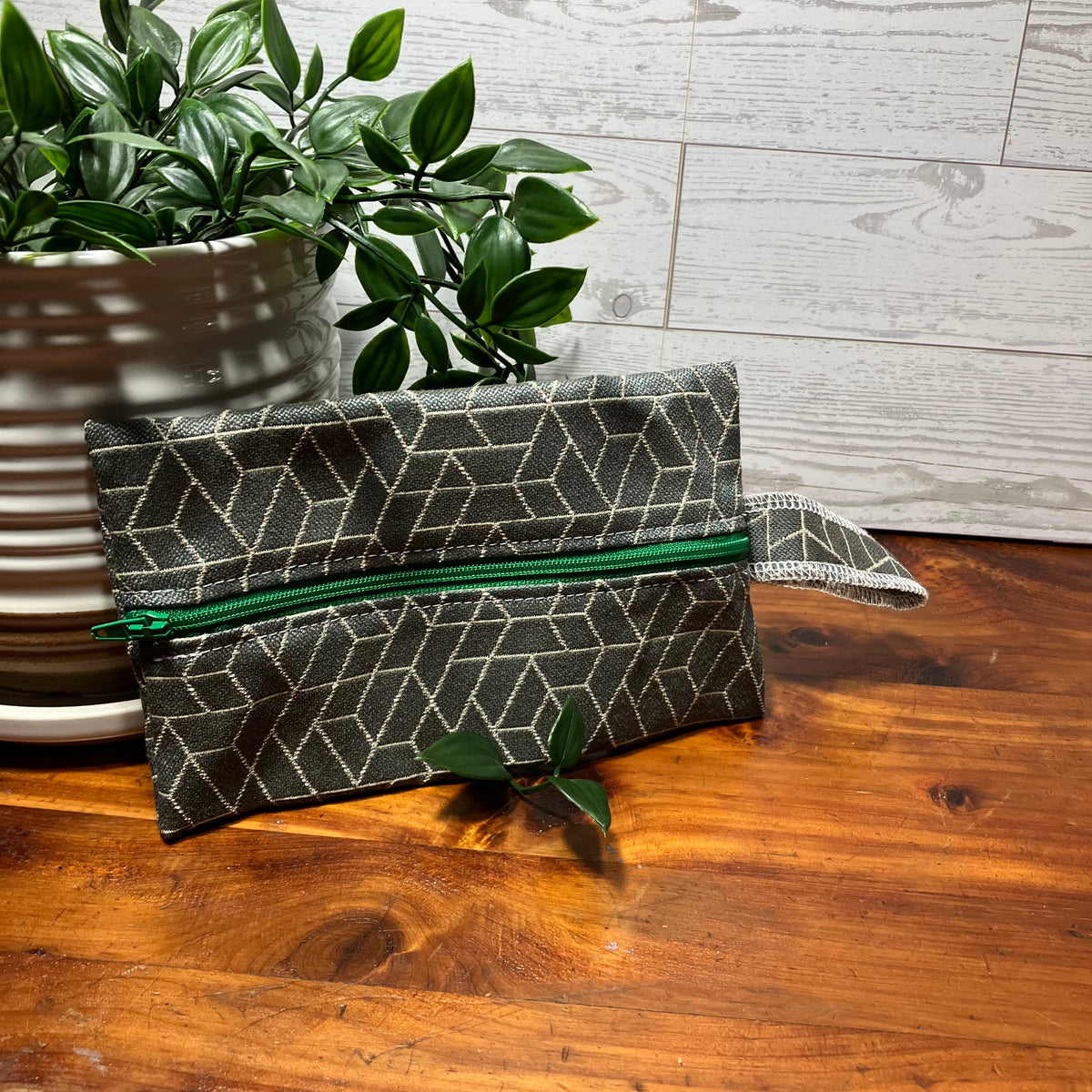 A small zip pouch with a dark gray and beige geometric pattern and a green zipper, resting on a wooden table next to a potted green plant.