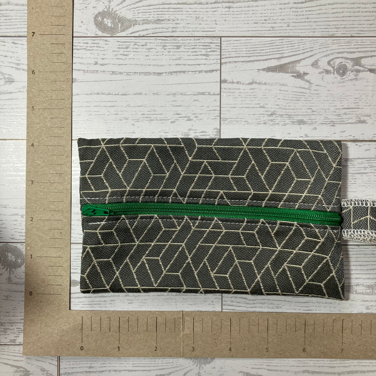 A small zippered pouch with a dark gray and beige geometric pattern and a bright green zipper, shown on a light wood background with a cardboard ruler for scale.