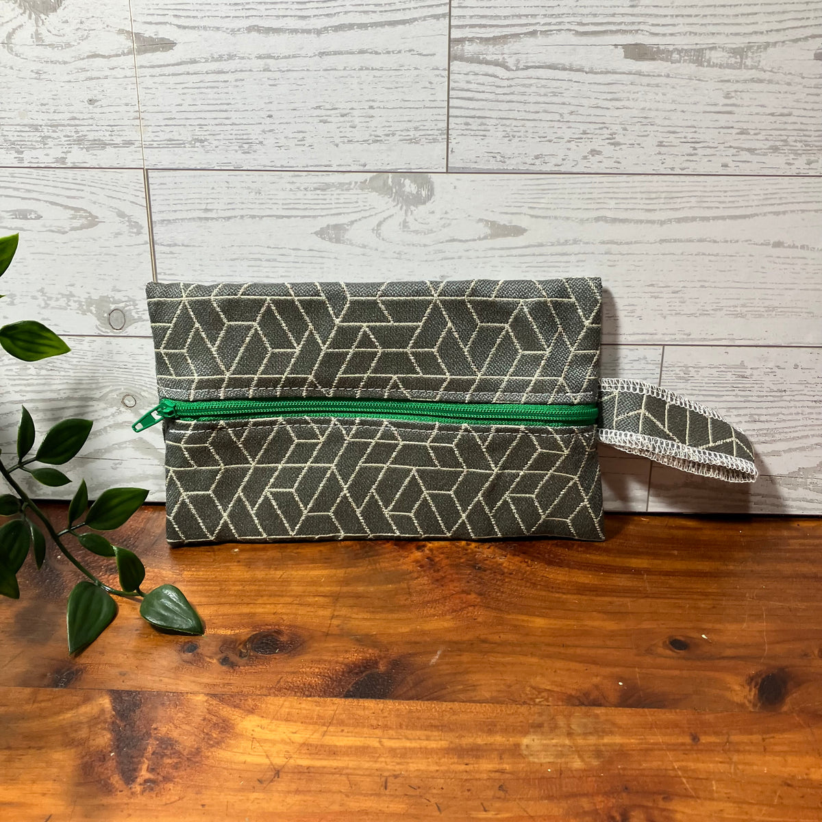 A fabric zip pouch with a geometric pattern in dark green and beige, featuring a bright green zipper and a matching wrist strap, displayed on a wooden surface against a light wood paneled background.