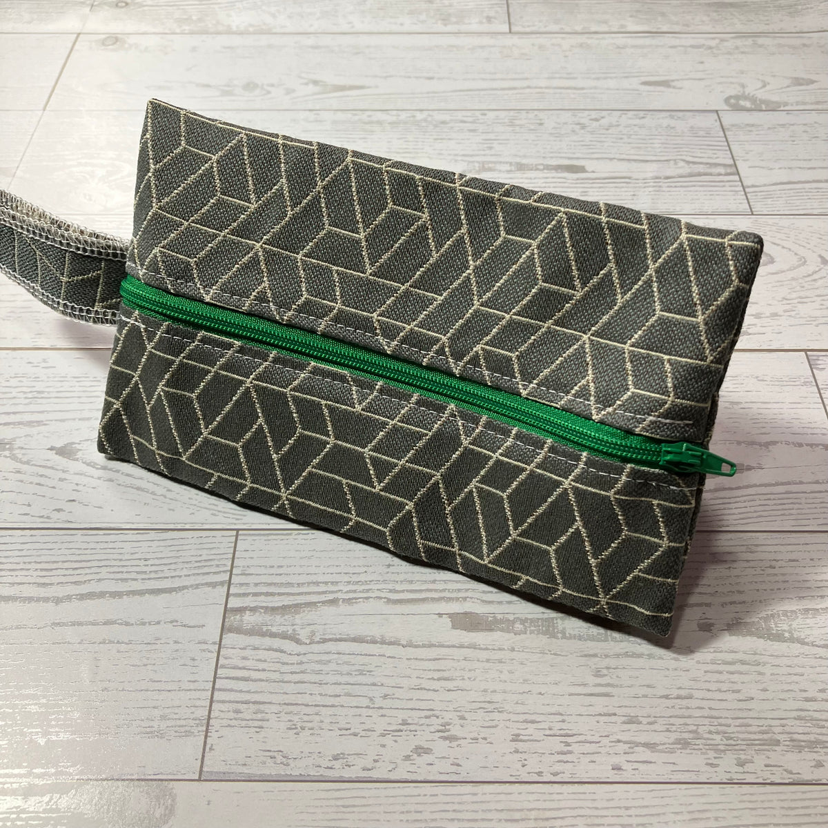 A rectangular zippered pouch with a geometric pattern in dark gray and beige, featuring a bright green zipper, resting on a light gray background.